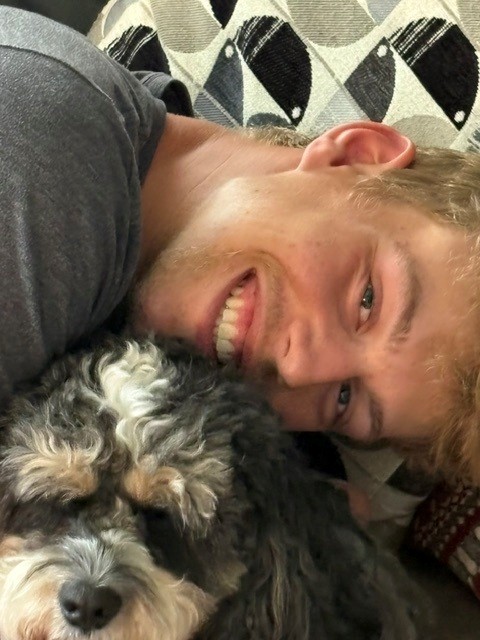 Adam Darveaux lying on a couch, smiling, with his dog resting against his cheek.